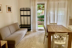 Une pièce avec un canapé beige, une table à manger en bois et des chaises blanches, un banc en métal noir, et une vue sur un jardin par une porte vitrée.