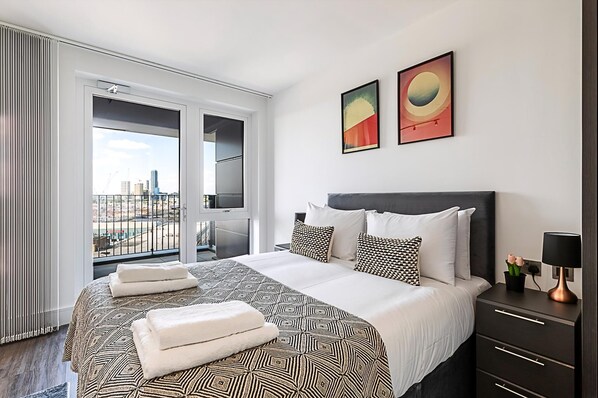 A modern hotel room with a large bed, a nightstand, and a view of the city through a balcony. - Sky-high Apartment With Modern Decor (London)