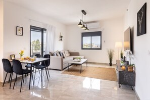 A modern living room with a dining area, a sofa, and a coffee table. There is a large window providing a view of the outdoors.