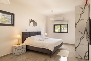 A bedroom with a bed, nightstand, lamp, artwork, window, and air conditioning unit. - Mamad Port Views Vista apartment (Haifa)