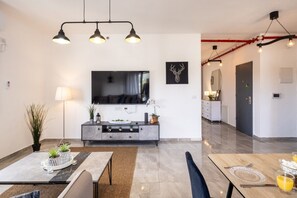 A modern living room with a flat-screen TV, a dining area with a table set for two, and a kitchen in the background.