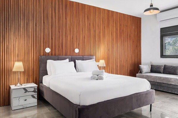 A modern bedroom with a large bed, two bedside lamps, a nightstand, and a grey sofa.