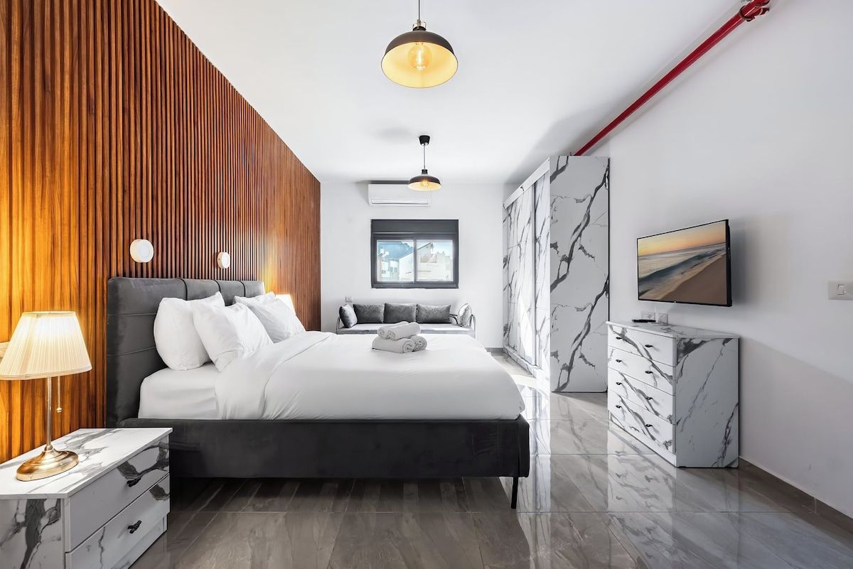 Een moderne slaapkamer met een bed, nachtkastje, lamp en een televisie aan de muur.
