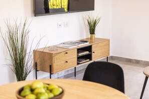 A modern living room with a wooden cabinet, a flat-screen TV mounted on the wall, and a dining table with a bowl of fruit. - Cozy 3BR Apt • Baháʼí Gardens by SeaNRent (Haifa)