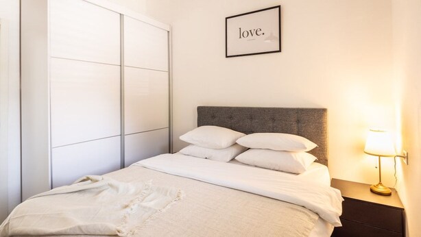Un lit bien fait, avec une tête de lit grise et du linge de lit blanc, une lampe de chevet et un cadre contenant le mot « amour ».