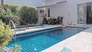 En modern utomhuspool med ett vackert, klart blått vattenområde, omgiven av en vit byggnad med en glasdörr och en överbyggd sittplats.