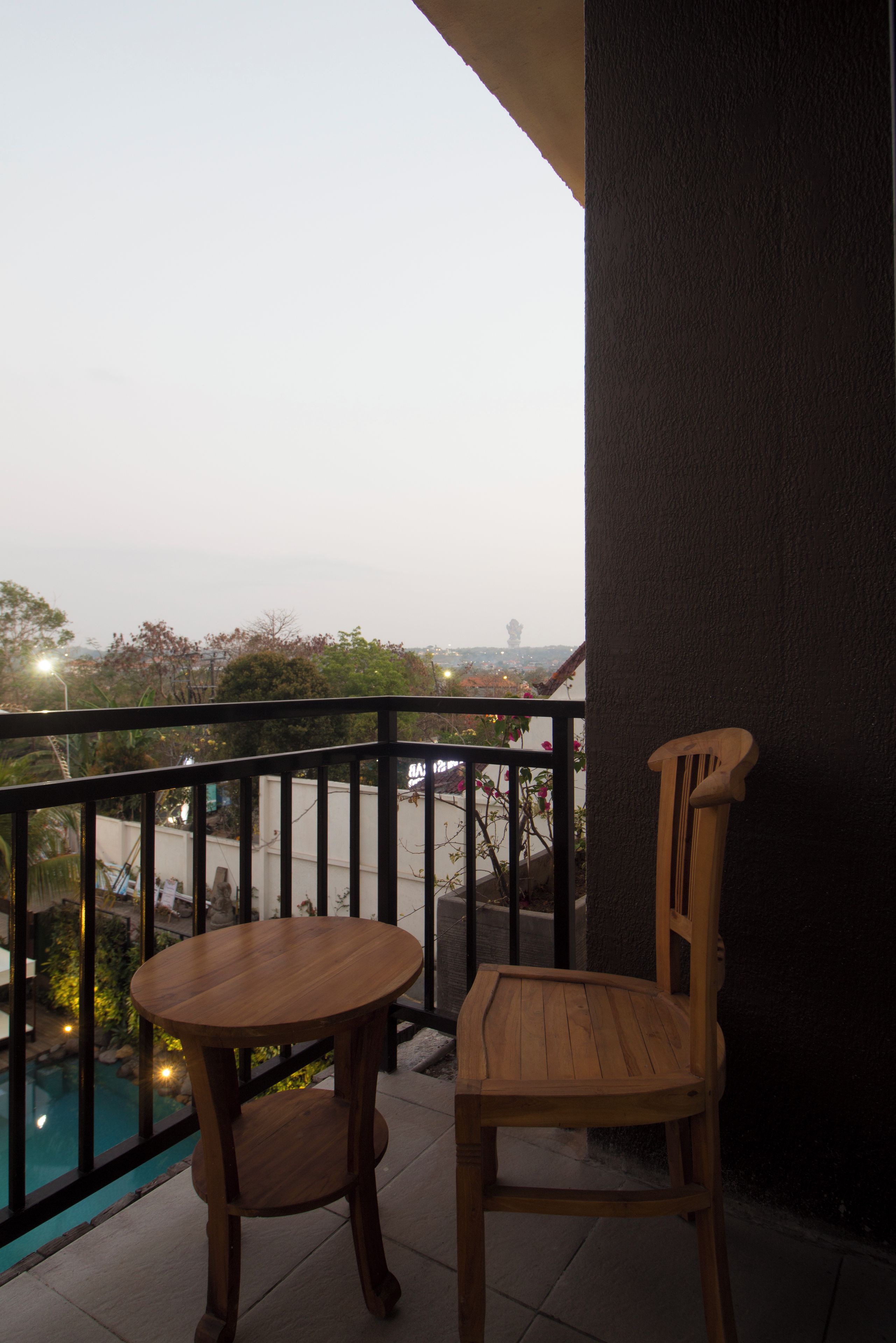 A balcony with a wooden table and chair, overlooking a swimming pool and distant cityscape.
