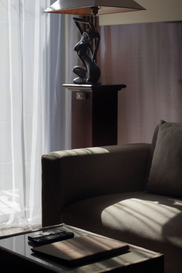 A room with a sofa, a lamp, a remote control, and a window with sheer curtains.