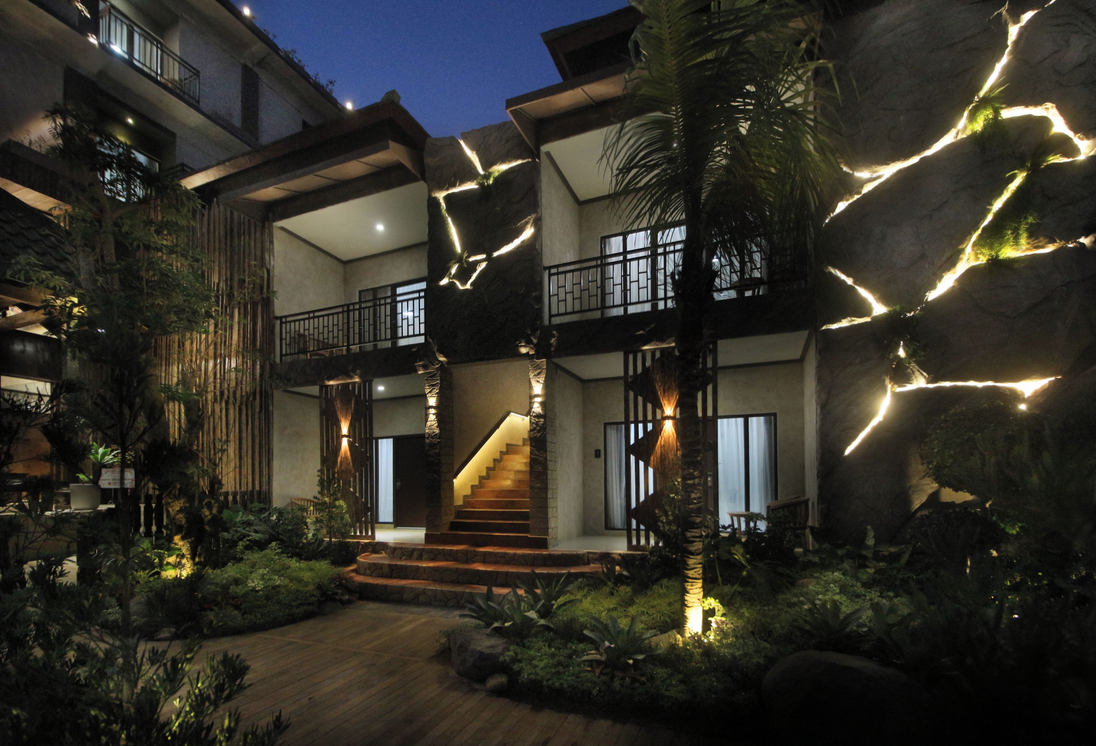 Un edificio de varios niveles con una escalera central, rodeado de vegetación e iluminado con luces colgantes.