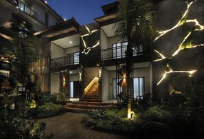 Un edificio de varios niveles con una escalera central, rodeado de vegetación e iluminado con luces colgantes.