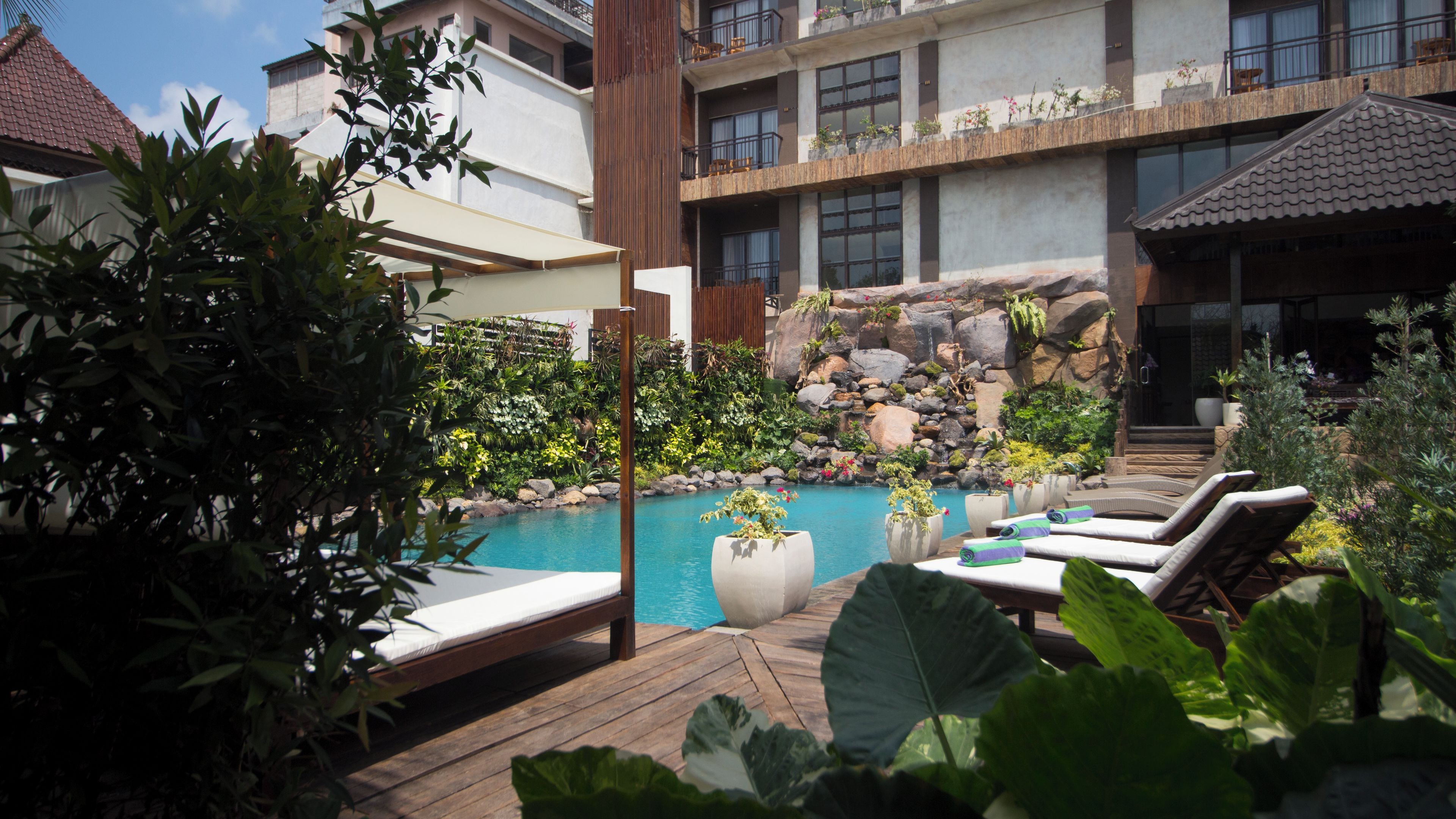 Un área de piscina con sillas de descanso, una terraza de madera y diversos tipos de plantas y rocas.