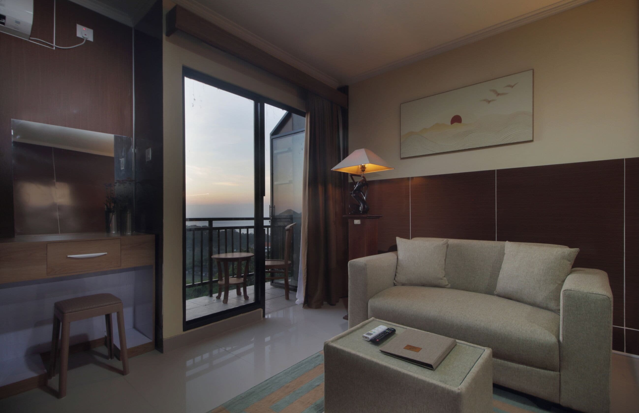 Quarto de hotel com sofá, uma mesa pequena com um controle remoto, abajur, quadro na parede e varanda com vista.