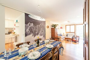 A dining area with a table set for four, a kitchen in the background, and a living room with a sofa and a window.