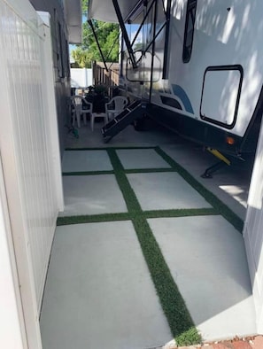 Een overdekt buitengebied met een betonnen vloer, een klein terras met een tafel en stoelen, en een witte camper in de buurt.