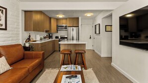 Eine moderne Küche mit Holzausstattung, einem Edelstahlkühlschrank und einer Mikrowelle. Ein Barhocker und ein Holztisch mit Zeitschriften sind ebenfalls vorhanden.