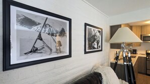 Ein Wohnzimmer mit einer weißen Ziegelwand, zwei gerahmten Schwarzweißfotos, einer weichen, weißen Decke auf einer dunklen Couch und einer Stativlampe.
