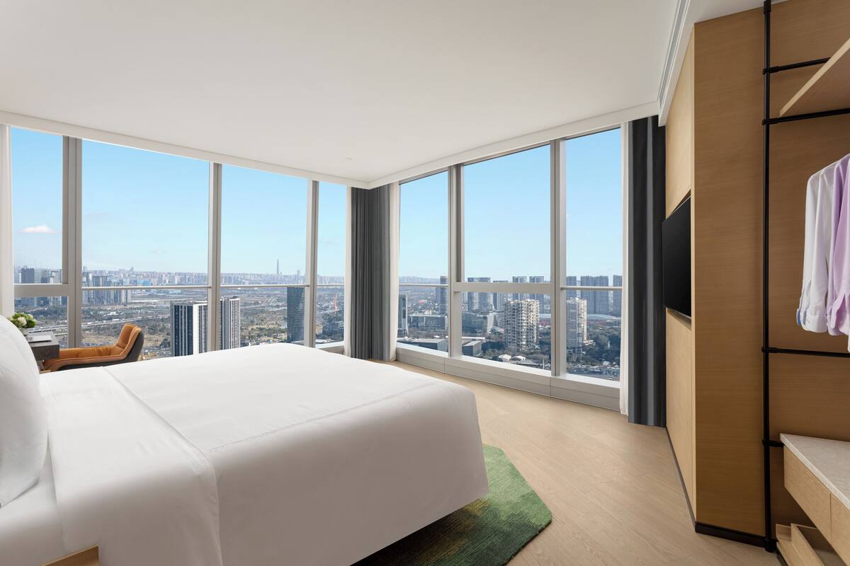 A modern hotel room with a large bed, a city view, a television, and a wooden wardrobe.