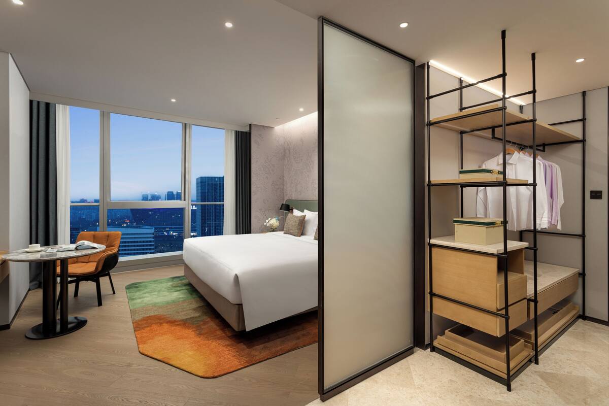 A modern hotel room with a large bed, a desk, and a view of the cityscape.