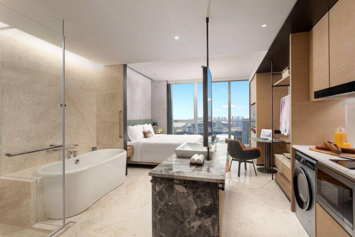 A modern hotel room with a large bed, a bathroom with a bathtub, a kitchen area, and a view of the city.