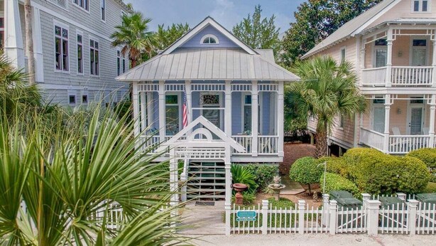 A coastal house with a white picket fence, a small porch, and a garden with palm trees.