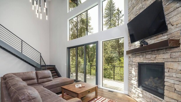 Una sala de estar moderna con un sofá seccional, una chimenea, un televisor montado en una pared de piedra y una escalera que conduce a un balcón con ventanas grandes.