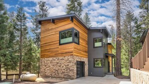 Una casa moderna de dos pisos con fachada de piedra y paneles de madera, rodeada de árboles altos.