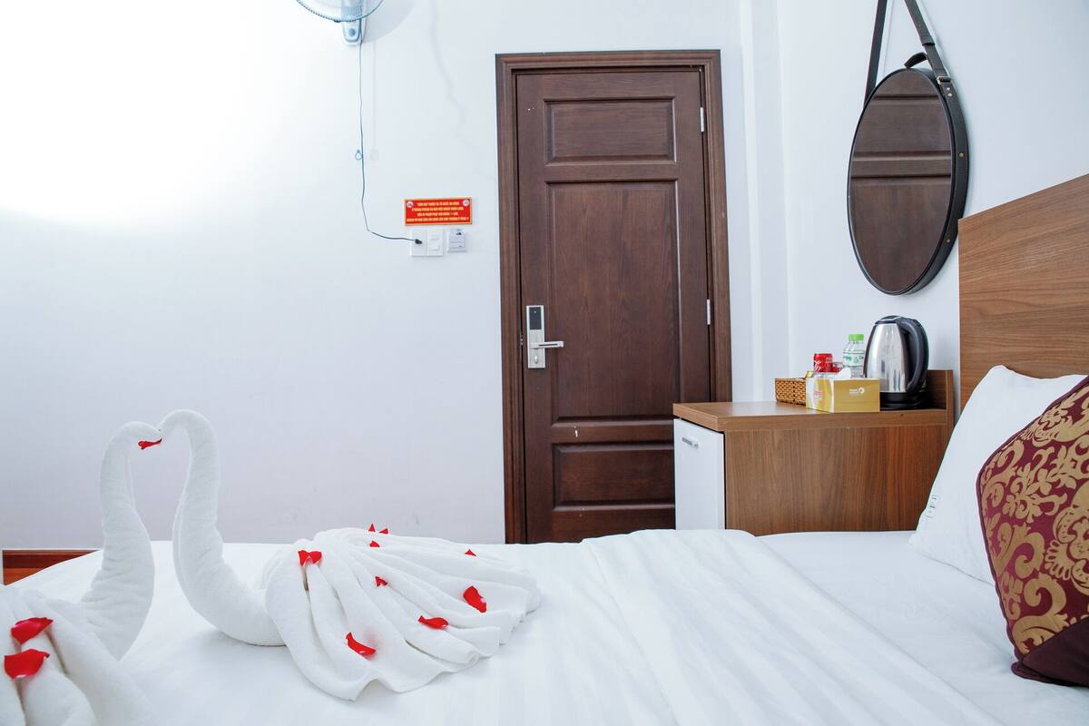 A hotel room with a bed, a wooden nightstand, a mirror, a door, and a wall-mounted fan.