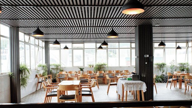 Un amplio comedor interior con mesas y sillas de madera, ventanas grandes y lámparas colgantes.