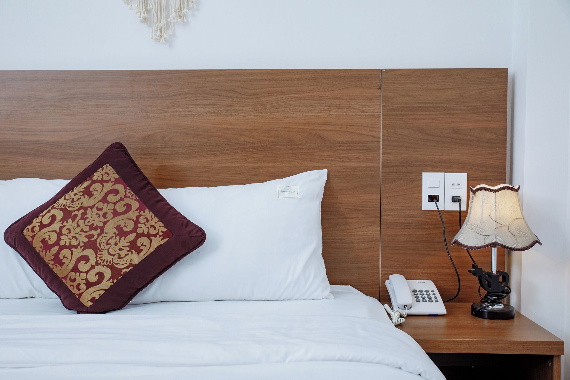 A neatly made bed with white linens and a decorative red and gold pillow. A bedside table holds a lamp and a telephone.