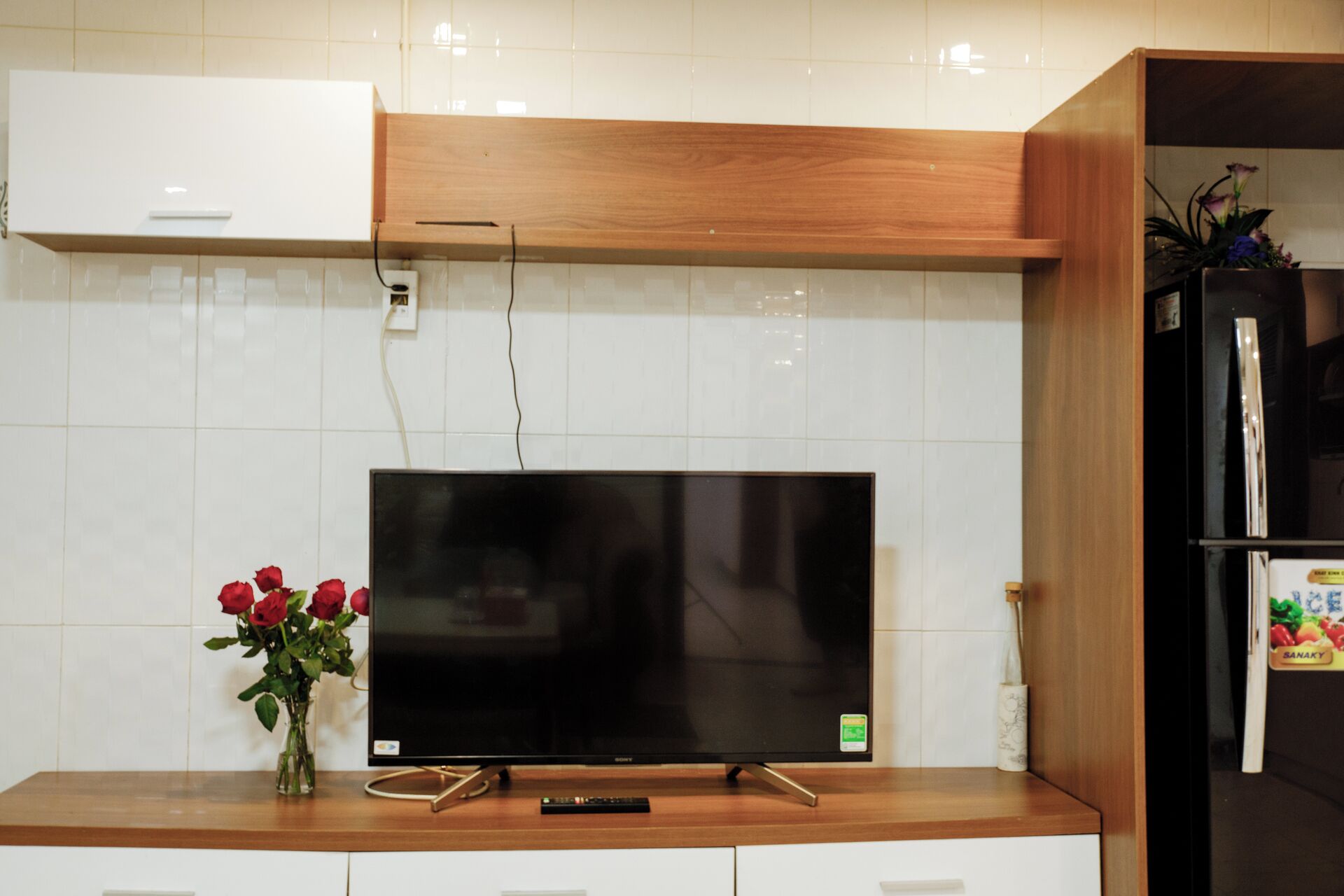 A kitchen with a flat-screen TV on a wooden counter, a vase with red roses, and a black refrigerator.