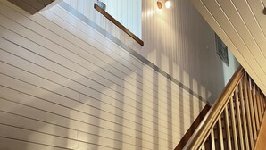 Un escalier en bois avec une rampe, menant à une pièce aux murs recouverts de panneaux blancs et dotée d’un puits de lumière.
