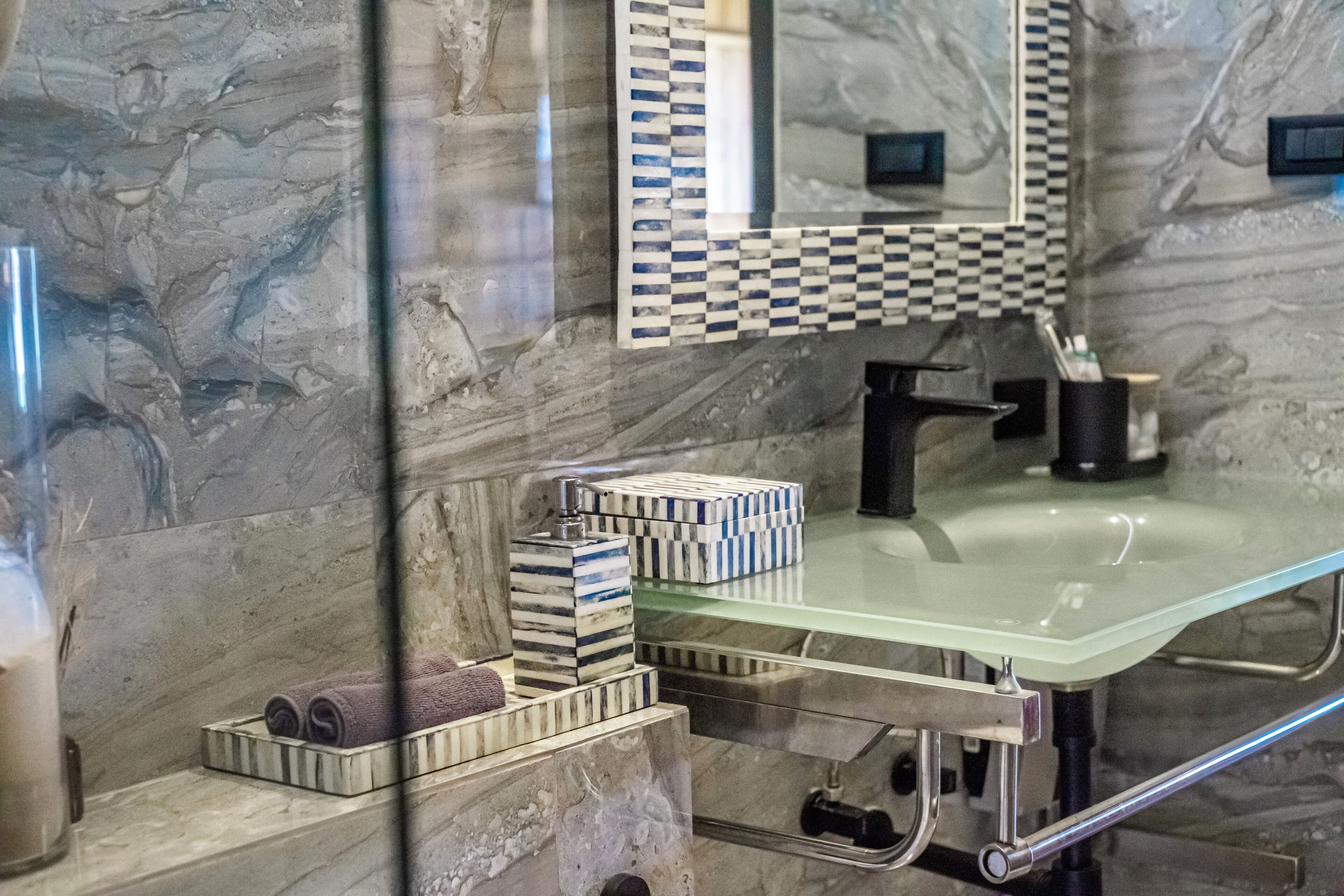 A bathroom with a glass sink, marble walls, a checkered mirror, and a towel rack with neatly rolled towels.