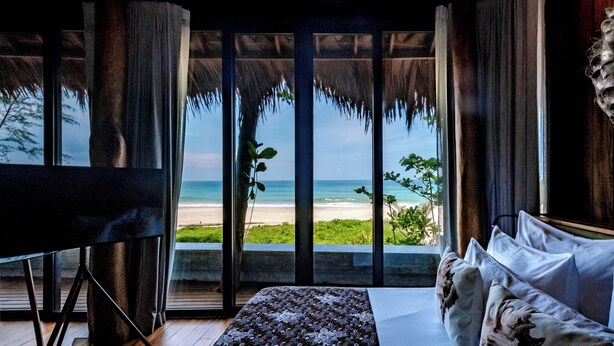 A room with a large window overlooking a beach, a bed with floral pillows, and a wooden floor. - Sealord Naithon Beach Phuket (Sa Khu)
