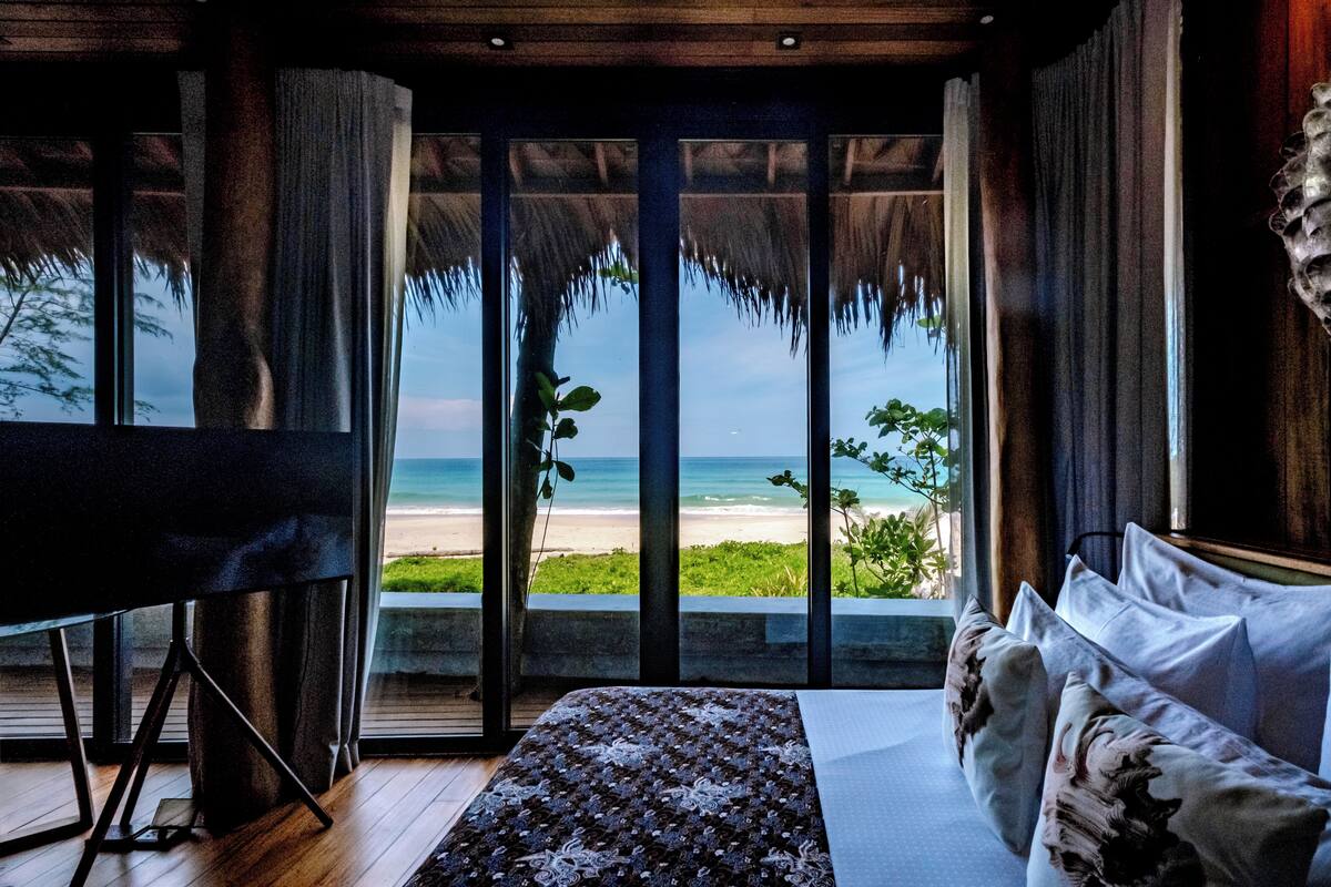 A room with a large window overlooking a beach, a bed with floral pillows, and a wooden floor.