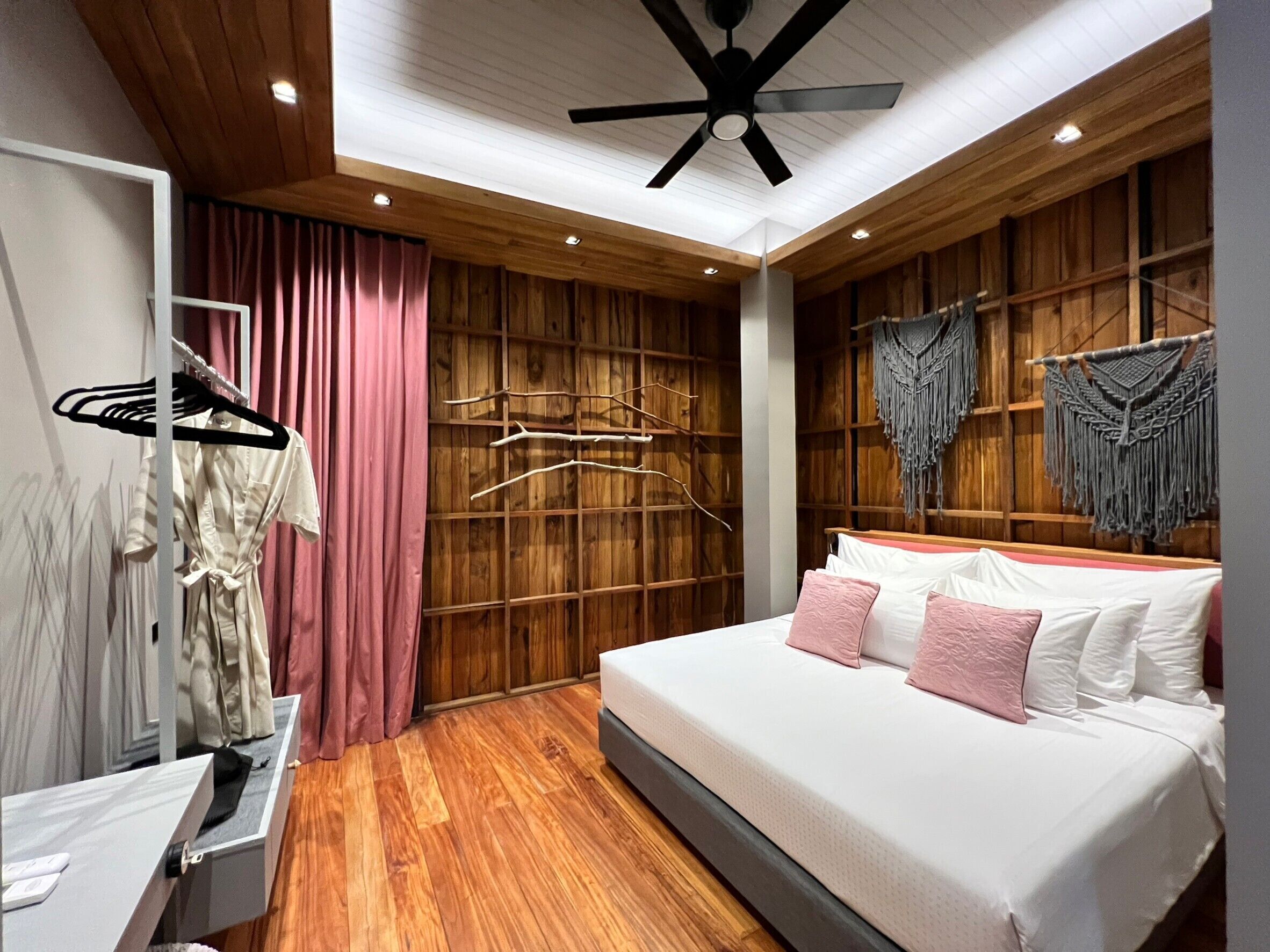 A modern bedroom with a wooden floor, a bed with white and pink bedding, a ceiling fan, and a wooden wardrobe with a curtain.