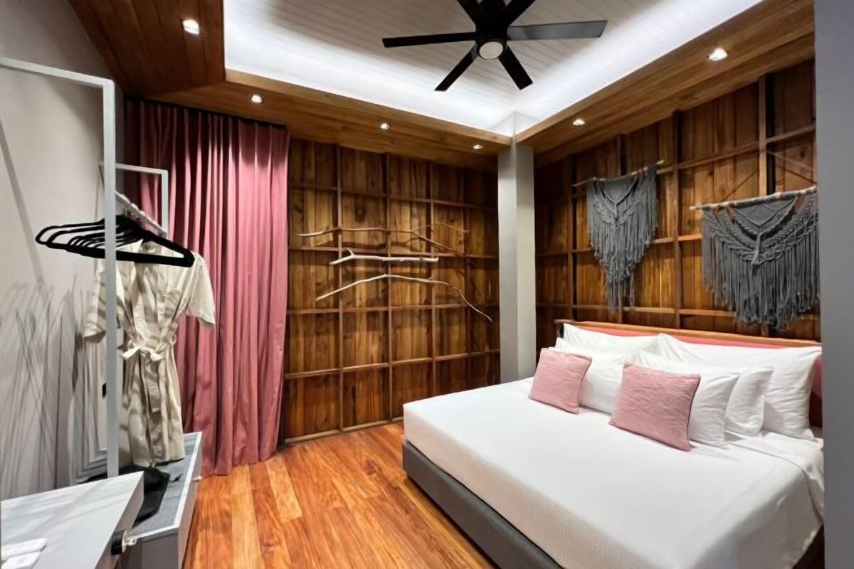 Un dormitorio con techo de madera, cama con ropa de cama blanca y rosa, armario y perchero con ropa.