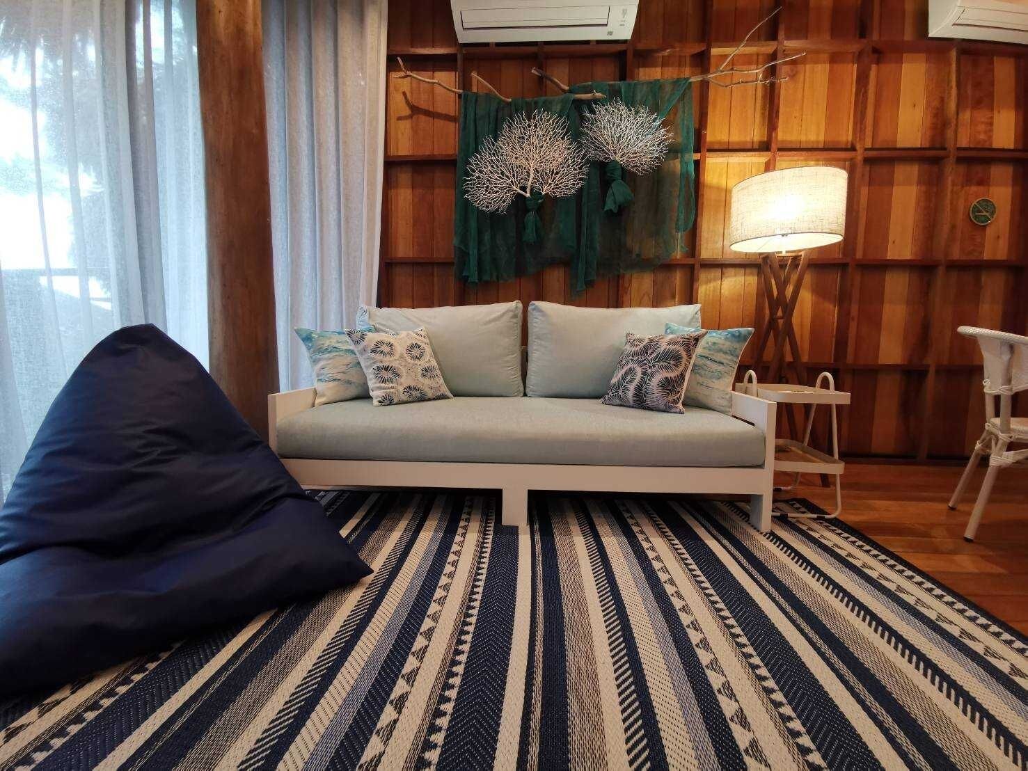 Un salon comprenant un canapé, des coussins, un fauteuil poire, un tapis à motifs, une lampe et un mur avec un lambris en bois et des éléments décoratifs.