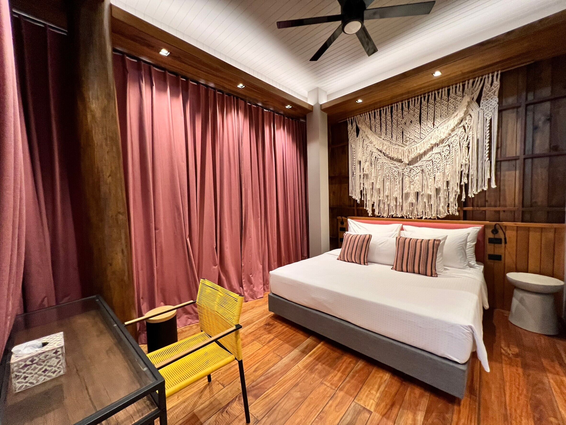 A bedroom with a large bed, wooden flooring, a ceiling fan, and a decorative macramé wall hanging.