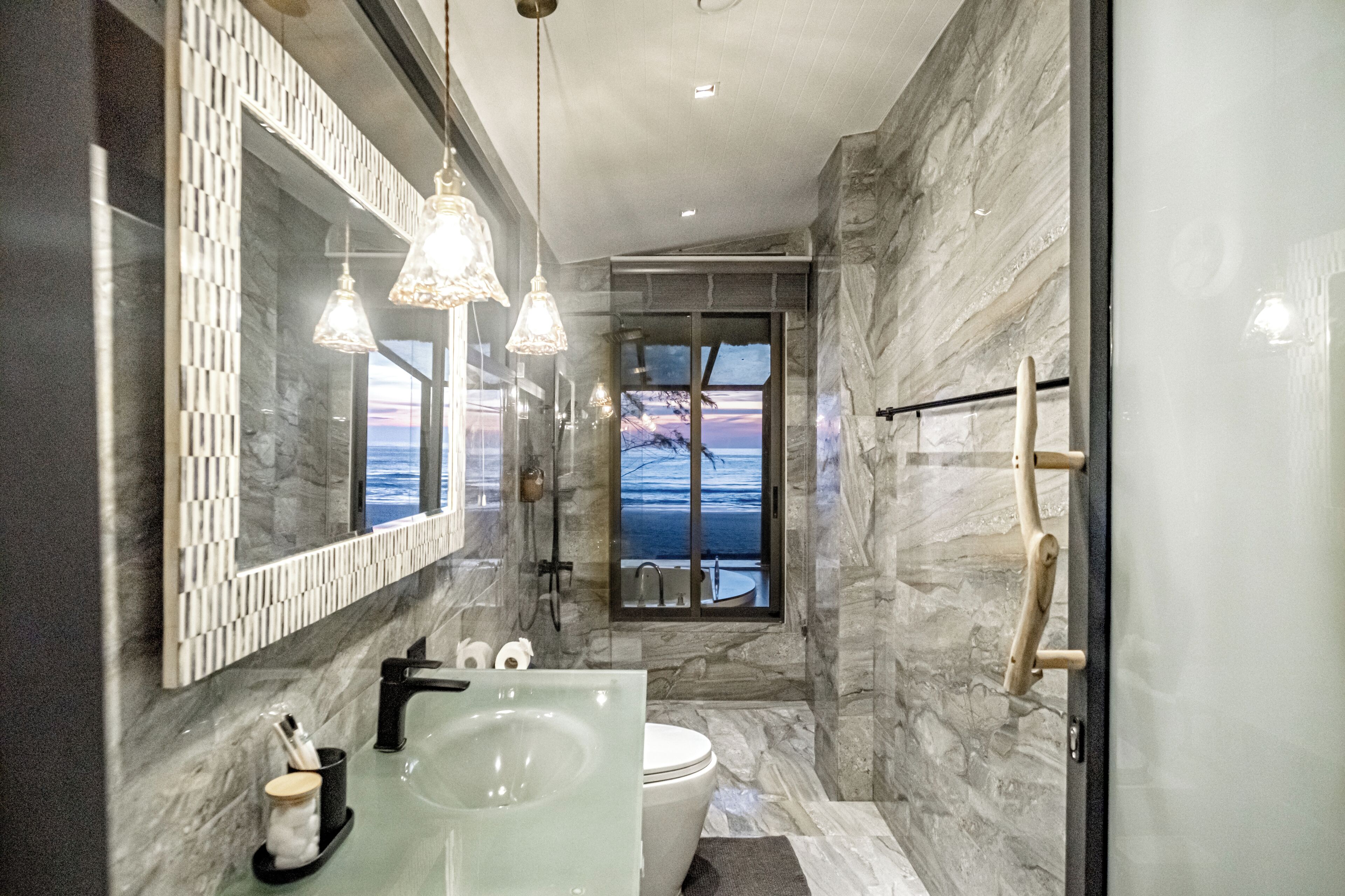 A modern bathroom with a glass-enclosed shower, a marble wall, and a view of the ocean.