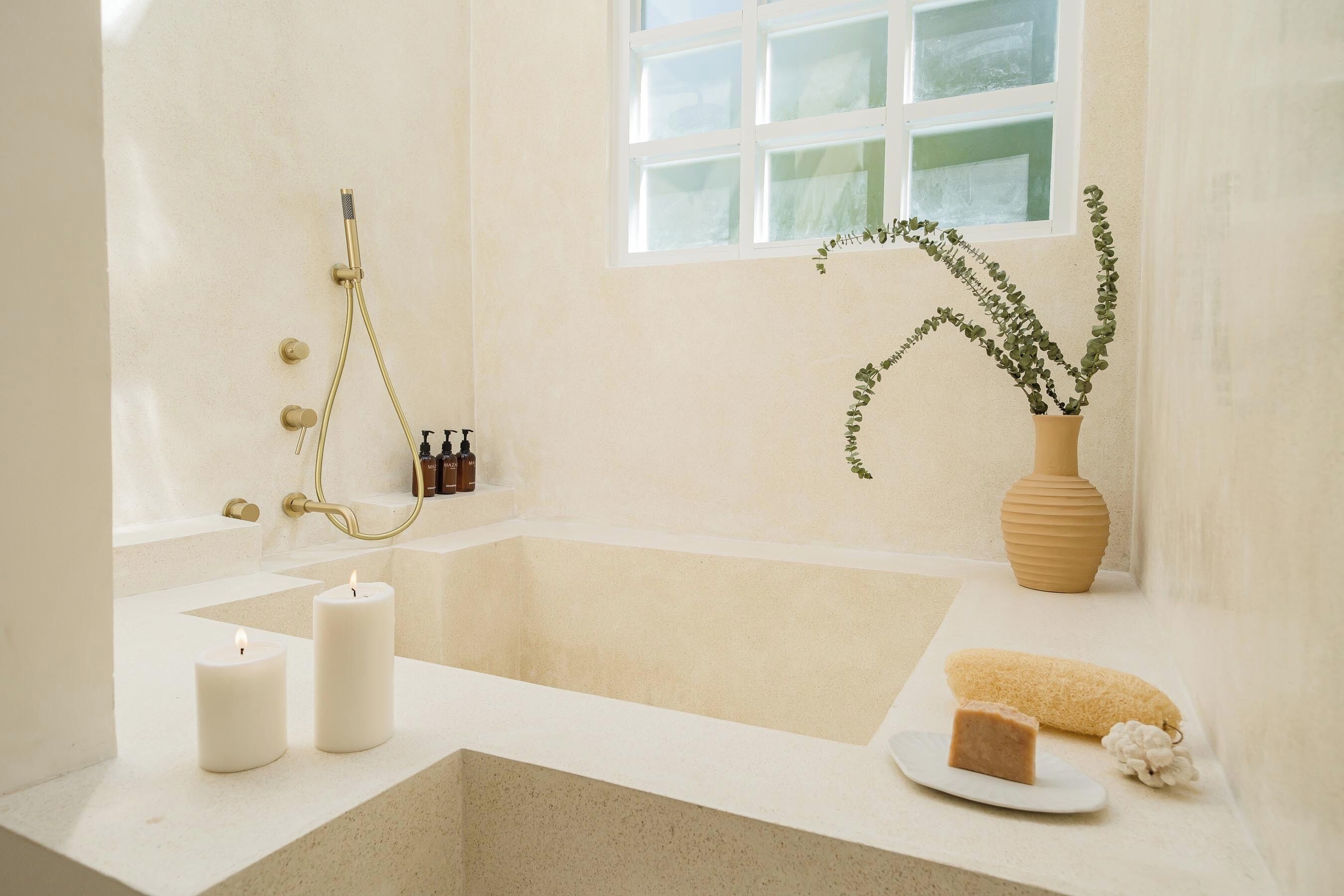A bathroom with a bathtub, two candles, a plant, and toiletries on the counter.