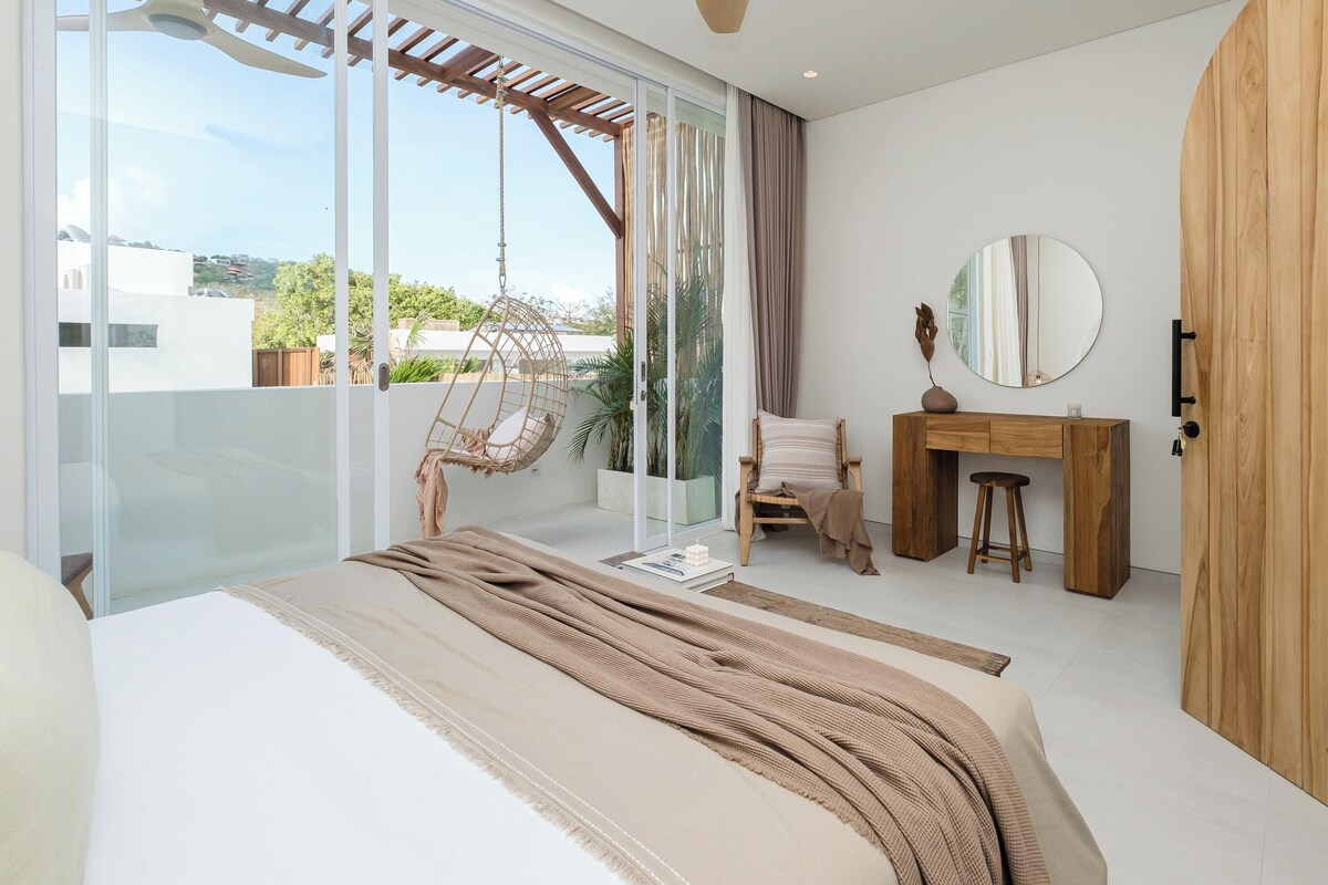 Un dormitorio moderno con cama, un escritorio de madera, una silla, una silla colgante y un ventanal con vistas a edificios y árboles.