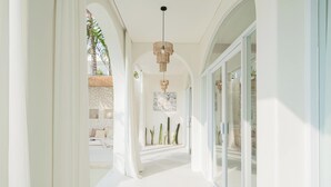 Un pasillo luminoso con paredes blancas, una lámpara colgante y vistas a una habitación con cama y plantas.