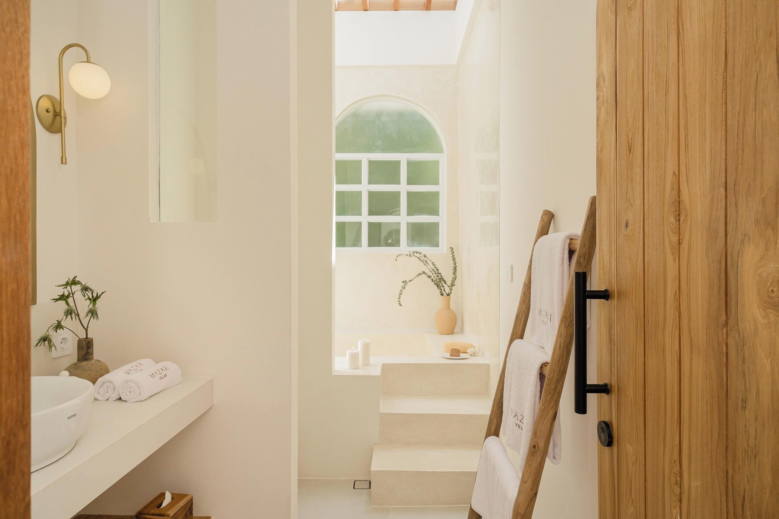Ein Badezimmer mit weißem Waschbecken, einem Spiegel, einem Handtuchhalter, einer Holztür und einem Fenster mit Blick nach außen.