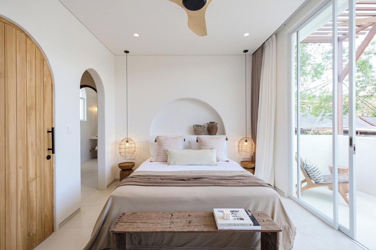A bedroom with a large bed, wooden wardrobe, arched doorway, and a view of the outside.