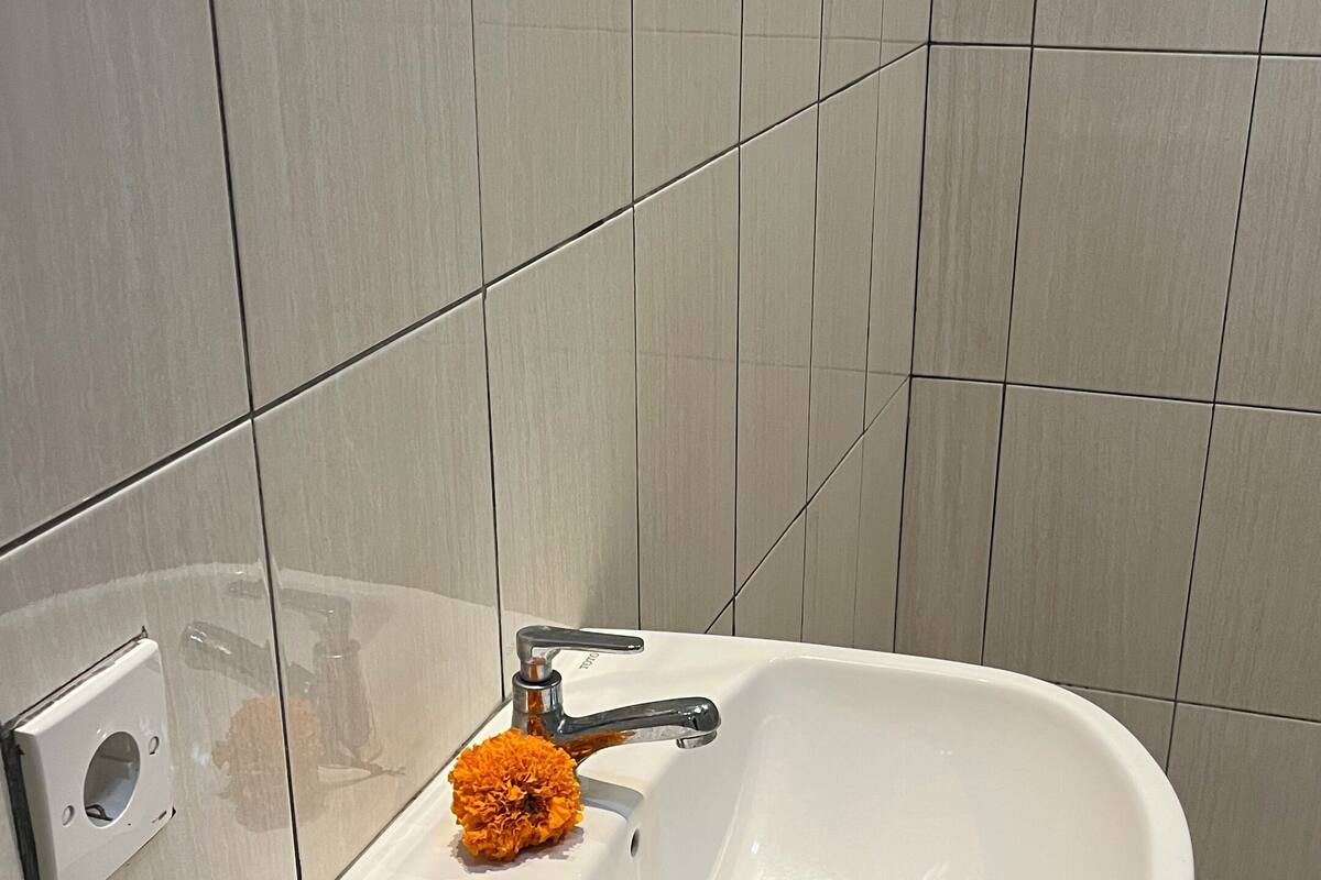 A bathroom with a white sink, a black trash bin, and a single orange flower on the sink.