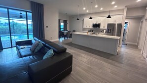 Una sala de estar moderna con un sofá de cuero negro, una zona de comedor con mesa y sillas, una cocina con armarios blancos y electrodomésticos de acero inoxidable, y un amplio ventanal corredero que da a un balcón con vista a una piscina.