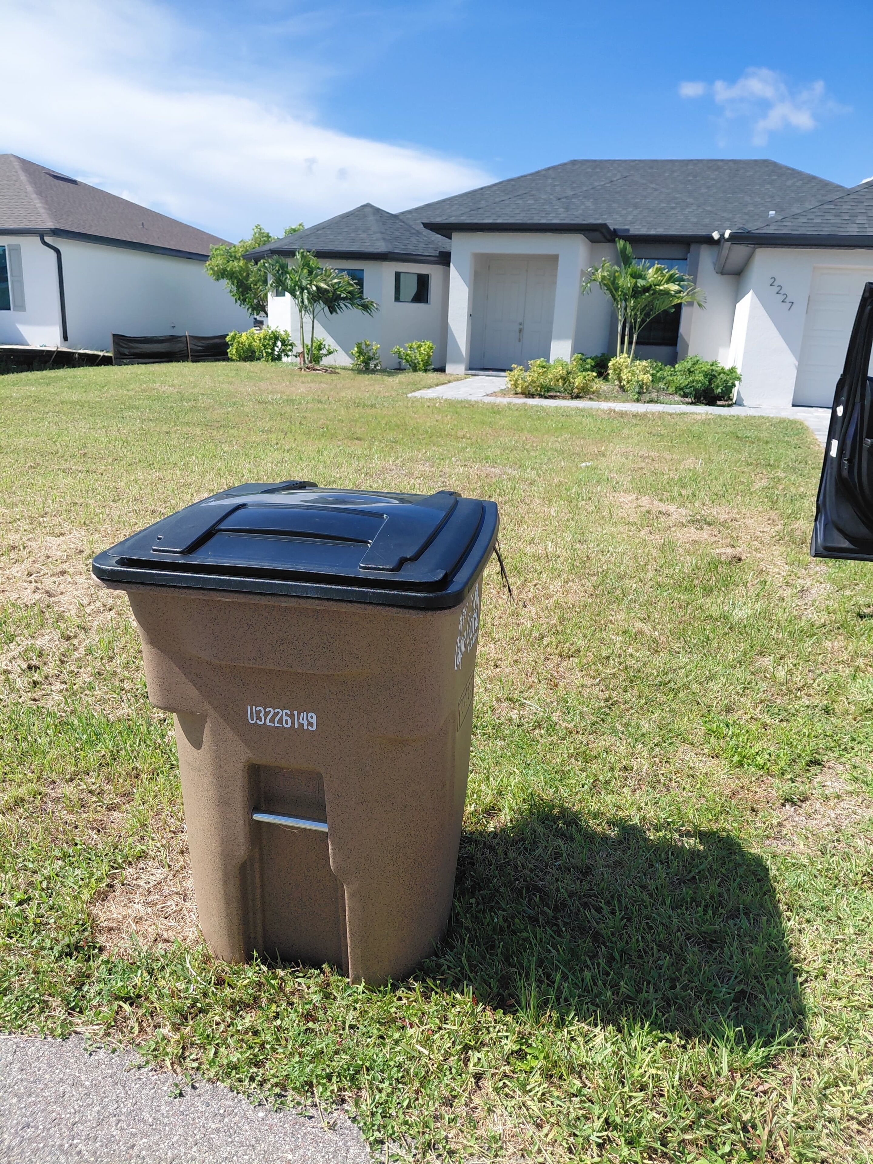A brown trash bin with a lid in a grassy area with houses in the background.