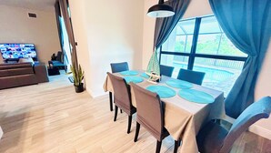 Un comedor con una mesa dispuesta para cuatro personas, un televisor al fondo y vista a una piscina a través de amplios ventanales.