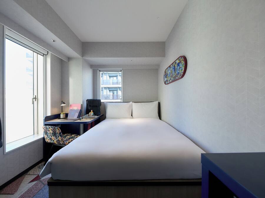 A modern hotel room with a large bed, a desk with a laptop, a chair, and a window with a view of buildings.