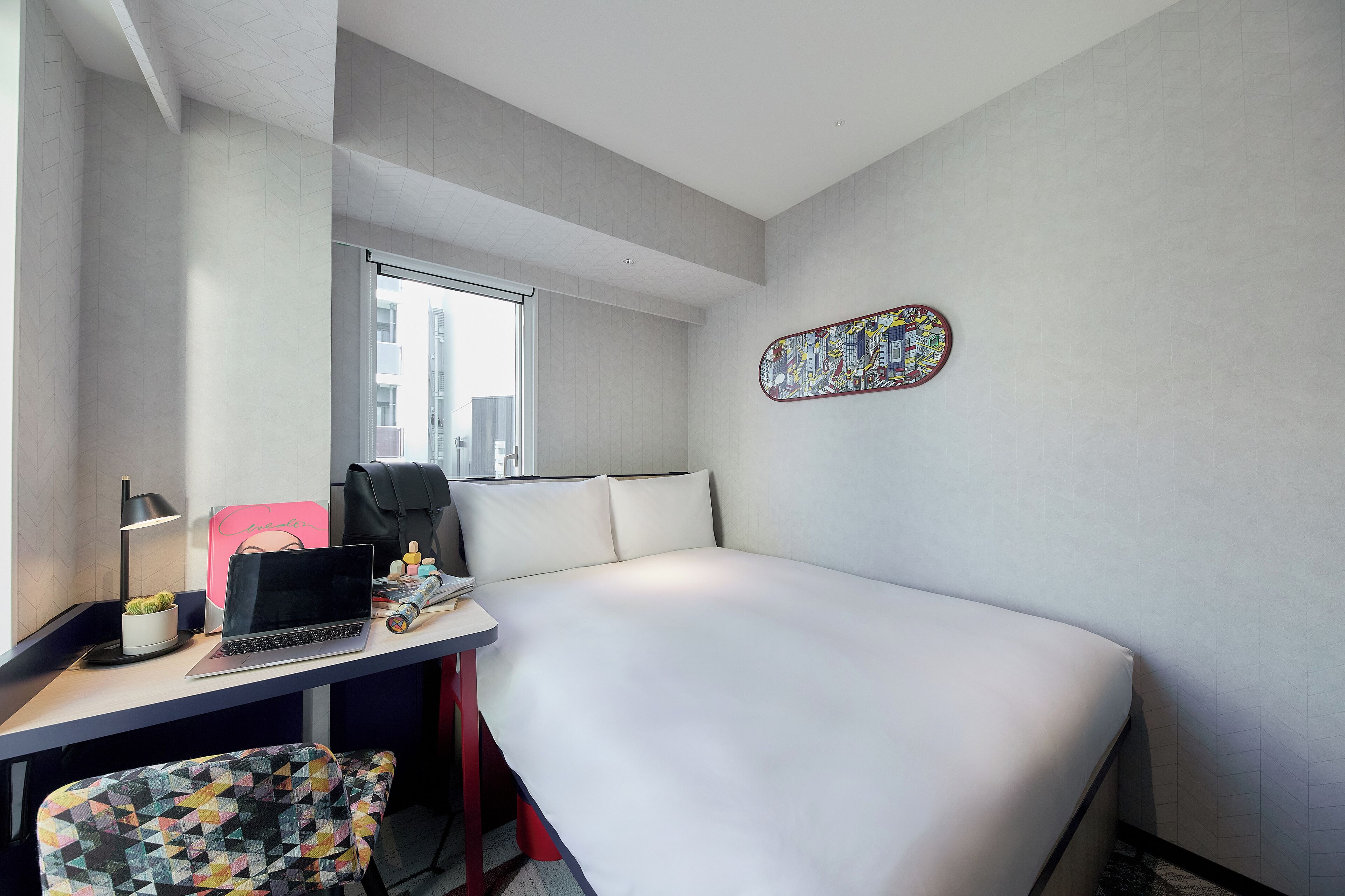 A hotel room with a bed, a desk with a laptop, a chair, a window, and a decorative wall piece.